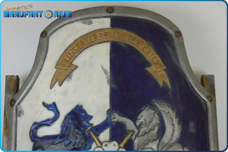 Detailansicht vom oberen Bereich des LARP Schild Löwe und Greif. Auf dem pergamentfarbigen Spruchband der erste Teil des Wahlspruchs "Unser Erbe Unser Krieg"